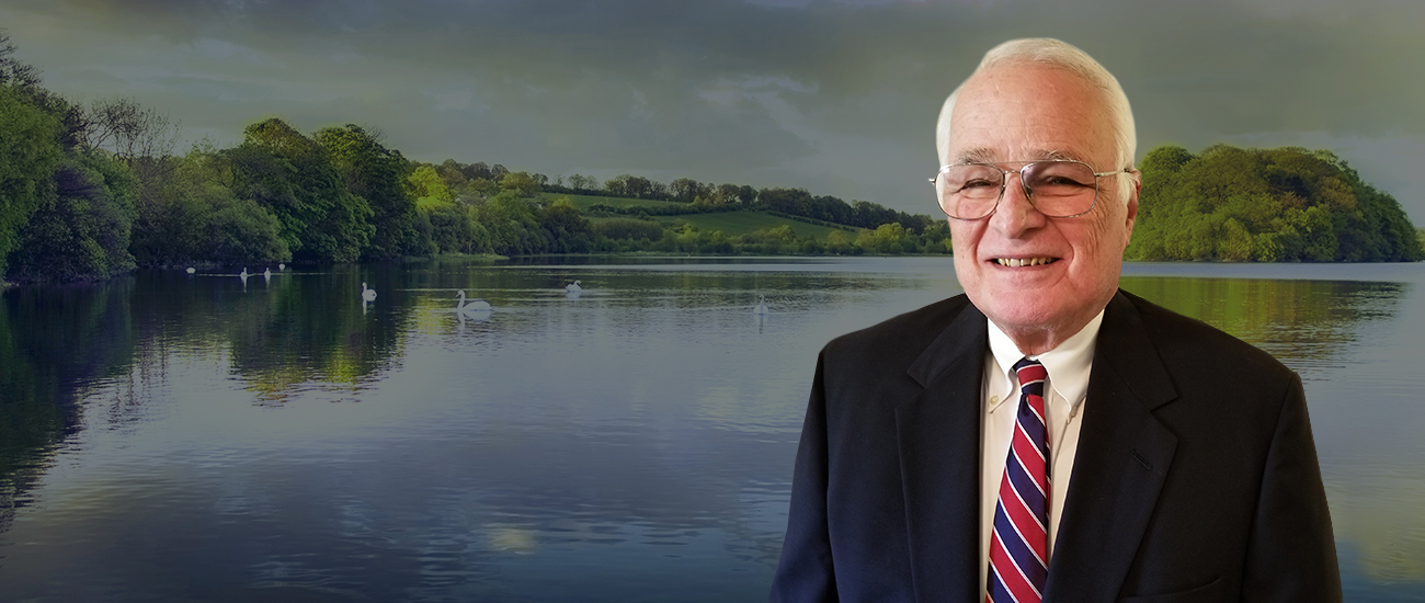 David S. Greenberg smiling in a suit and tie with a scenic lake and cloudy sky in the background.